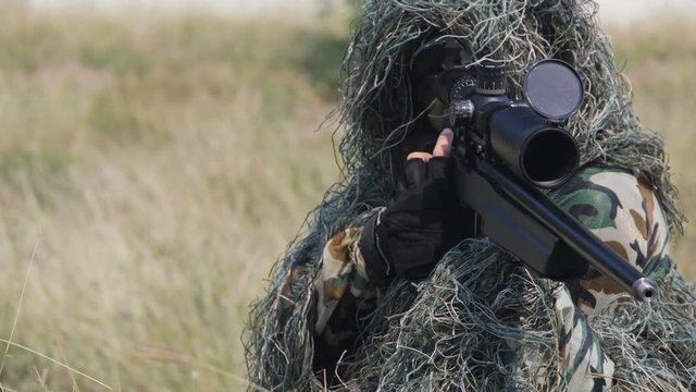 Army sniper wearing disguise camouflage suit sitting and aiming in grass carrying his gun