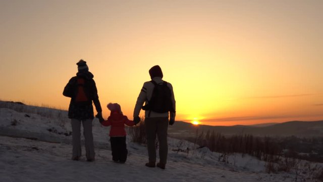 Happy family of three people standing on a mountain holding hands in winter at sunset.