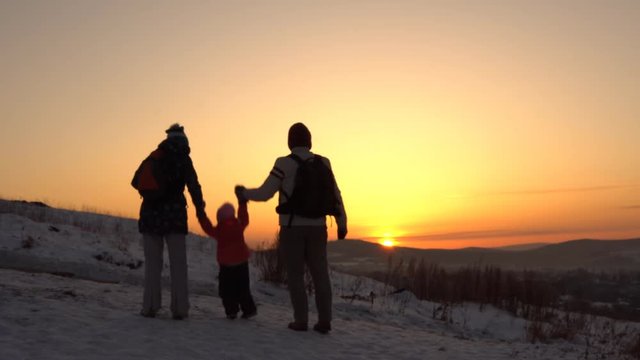Happy family of three people standing on a mountain holding hands in winter at sunset. Parents raise the child up.