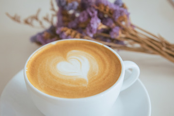 a heart pattern late-art coffee in a white clean cup with a purple bouquet flower on table