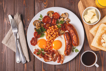 Traditional full English breakfast with fried eggs, sausages, beans, mushrooms, grilled tomatoes and bacon on wooden background.