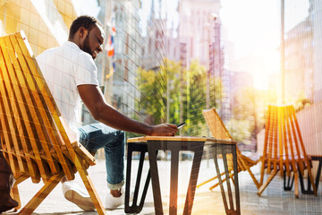 Pleasant afternoon. Handsome young positive man sitting in a comfortable chair in a lovely cafe and smiling while looking at the screen of his phone and waiting for an important message