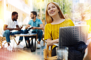 Noticing friends. Pretty smiling young woman sitting in a cafe with a laptop on her knees and feeling happy while noticing her best friend in a crowd
