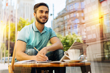 Wonderful day. Cheerful smiling handsome young man feeling happy while sitting in a quiet place and looking into the distance before making notes for his next lesson