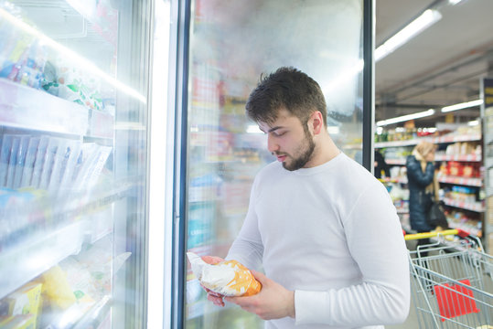 The Man Looks At The Label Of The Frozen Product Near The Fridge In The Supermarket. The Choice Of Frozen Foods In The Supermarket.