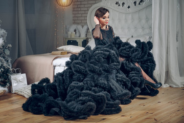 Beautiful woman in black evening dress sits on the bed in luxury interior. Fashion shot. the girl is going to a party.