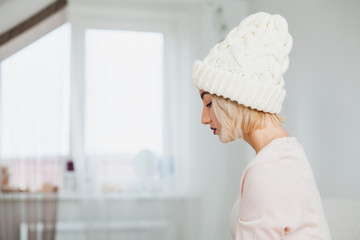 Portrait of young woman in white hand knitted hat. Profile of a girl indoors in warm clothes. Copy space for text. Hygge, comfort and home concept.