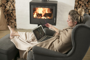 Beautiful middle-aged woman next to the fireplace relaxes in the living room and works on laptop from house. Girl in 30's surveyed for the  home bussiness project