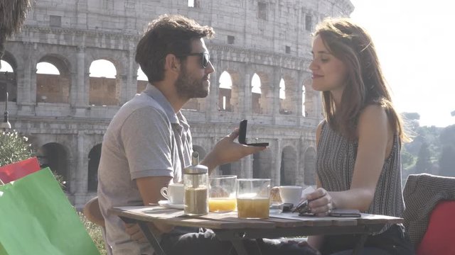 Happy couple marriage proposal with ring sitting at restaurant in front of colosseum in rome at sunset emotional FLAT NO GRADING