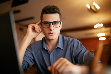 Authentic portrait of young confident businessman looking at camera with laptop in office. Hipster man in glasses and knitted sweater doing his startup ICO project. Smart casual wear. Business theme