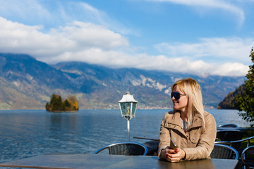 Fototapeta premium Young beautiful woman with mobile phone on the shore on the lake shore with mountains in a cafe. Communication and internet in travel concept