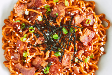 Close up Korean hot and spicy instant noodle in black and white bowl topped with bacon, sesame, seaweed and chopped scallion served with kimchi on wood table. Homemade delicious asian food concept.