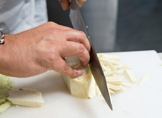 A knife cutting thin cubes of fresh organic celeriac root vegetable, on a white cutting board.