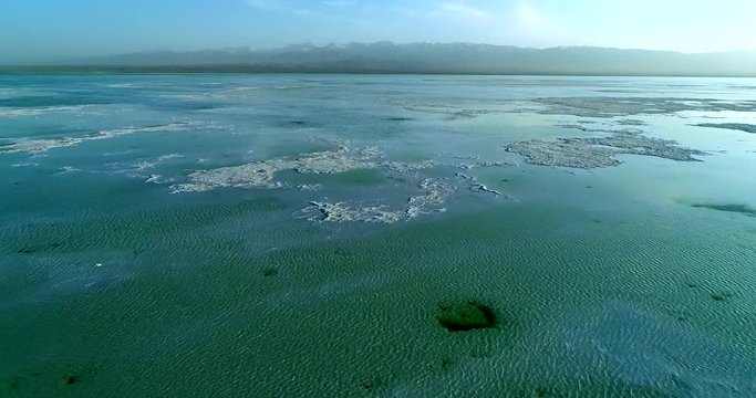 Flying drone over Chaka Salt Lake on the Tibetan Plateau showing the lake&rsquo;s thick salt bed that lies close to the water surface, a blue cloudy sky and mountains in the background. Part 1 of 2
