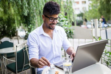 Young attractive afro American businessman sitting in cafe bar and doing some work on laptop. 