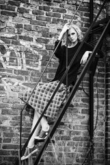 Grunge fashion: cute young girl (informal model) in checkered skirt and jacket sitting on ladder. Black and white
