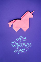 Handmade pink trendy geometrical origami unicorn on ultraviolet background. Are unicorns real text, letters, lettering, quote. Vertical poster, postcard, banner. © mykolastock