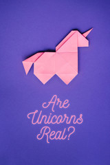 Handmade pink trendy geometrical origami unicorn on ultraviolet background. Are unicorns real text, letters, lettering, quote. Vertical poster, postcard, banner. © mykolastock