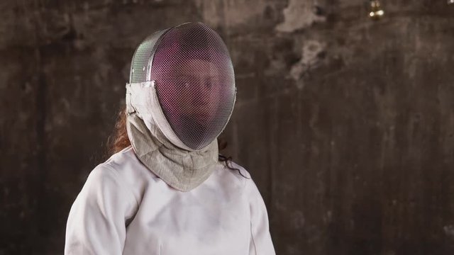 portrait of a young woman in a protective helmet, a lady stands against the background of an underground wall, a lady holding a rapier for sports competitions
