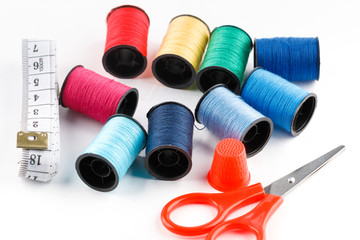 Red scissors, measuring tape and thimble with spools of thread on white background.