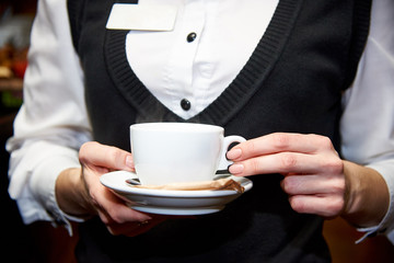A cup with a hot drink on a saucer in the hands of a waiter in uniform