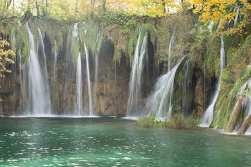 A long exposure of rushing water from various waterfalls in Plitvice lakes National Park. The trees are yellow and the mosses are light green and the water is turquoise.