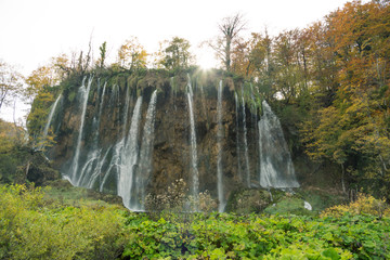 The sun shining above a waterfall in Plitvice Lakes National Park. The are lush green plants in the foreground and orange and yellow trees in the background above the waterfall. 