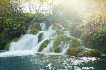 A sunny day looking at a small waterfall surrounded by lush green grass and turquoise waters. The background is a golden yellow. This is a long exposure.