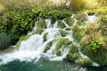 Light leak from the sun over a small waterfall in Plitvice Lakes national Park. The water is flowing in different directions and it passes the lush green grasses. The water is turquoise.