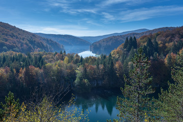 Two lakes are shown in Plitvice Lakes National Park. The trees are changing colors from green to yellow, orange and red. There is some fog above the lake and few clouds in the blue sky. Great distance