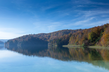 Reflection of a peninsula on a lake. Taken in Plitvice Lakes National Park. The sky is mostly clear with few clouds and the trees are turning colors.