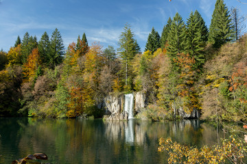 A waterfall and a lake in Croatia. The sky is mostly clear with few clouds and the trees are green and yellow. Taken at Plitvice Lakes National Park.