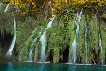 Long exposure of a small waterfall in Plitvice lakes National Park. The water is clear and turquoise colored. The trees above are golden and the moss is a lush green. 