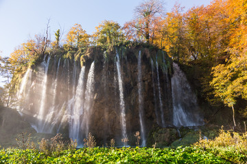 A giant waterfall with fog showing the sun rays beaming through. The picture was taken in the fall or autumn showing the changing colors of the trees.