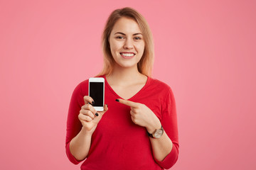 Cheerful blonde female with broad smile indicates at blank screen of smart phone for your advertising content, dressed in red sweater, isolated on pink background. Technology and people concept