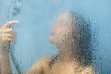 Female enjoys douching in shower cabine, poses naked against wet brurred glass background, takes...