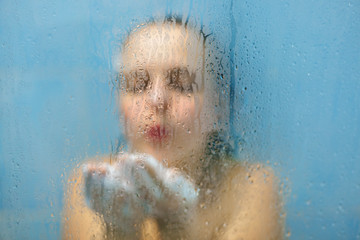 Restful young female blows foam on hands as stands in douche, has wet body, relaxes at bathroom alone, stands under hot water shower, washes her body, feels freshness and rejuvenation