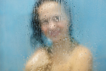 Glad female poses with nude perfect slender body in bathroom behind blurred sweat glass, showers in shower cabine, cares of skin, feels relaxed. Hygiene, freshness and natural beauty concept