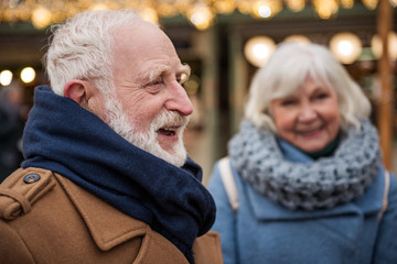 Side view profile of happy old man standing outdoor with relaxation and laughing. Senior woman is looking at him with joy on background