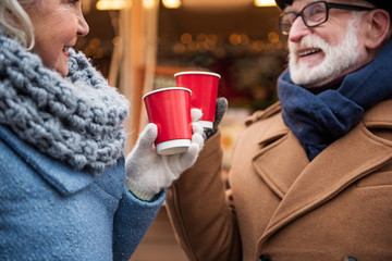 Close up of arms of cheerful old man and woman celebrating holiday outdoor. They are drinking mulled wine and laughing. Focus on red cups