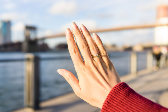 Young Woman's Hand With Diamond Engagement Ring Solitaire, Gold Wedding Band Outside Outdoors In NYC New York City Brooklyn Bridge Park By East River, Cityscape, Skyline Bokeh