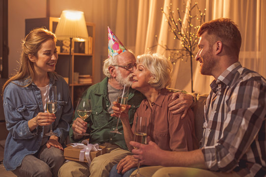 Smiling grandpa kissing cheerful grandmother in check. Outgoing relatives tasting alcohol on sofa. Love concept