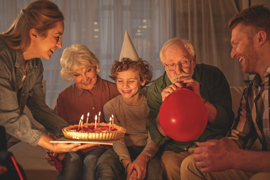 Outgoing mother giving cake with candles to cheerful son. Beaming grandmother sitting near him. Grandfather inflating balloon. Happy birthday concept