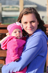 Obraz premium Close-up Portrait of Very Beautiful Teenage Girl with Long Brown Hair and Big Eyes looks at Camera Holding Her Sister baby Girl in Pink Outfit, Sunny Day, Oregon USA
