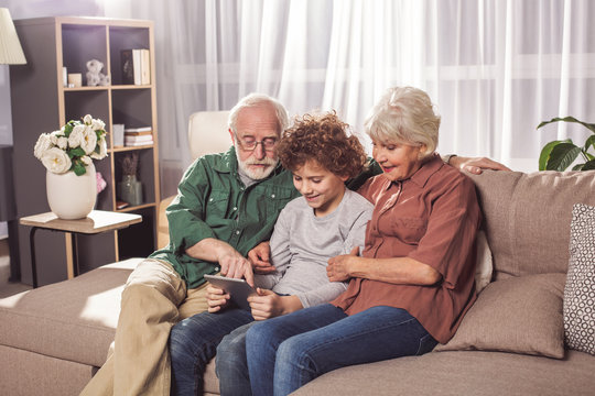 Portrait of smiling unshaven grandfather talking with happy kid and wife. They watching at electronic tablet while sitting on sofa. Entertainment concept