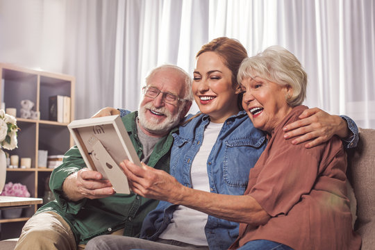 Portrait Of Laughing Grandparents And Smiling Young Woman Looking At Photo In Room. Remembrance Concept