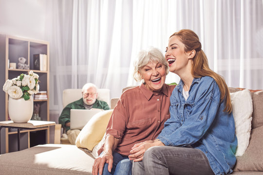 Portrait of cheerful old woman laughing with young lady on sofa. Beaming bearded grandfather typing in laptop. Happiness concept