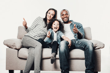 Portrait of content mother, father and daughter relaxing on sofa and showing thumbs. Isolated on background