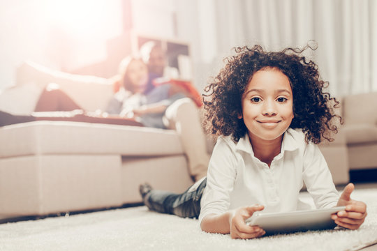 Portrait of enjoyed kid with device in hands. She is relaxing on the floor at home with comfort. Focus on girl. Parents on background
