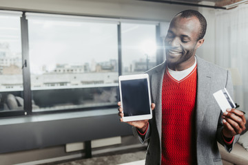 Waist up portrait of cheerful stylish businessman showing tablet and credit card indoors. Copy space in left side